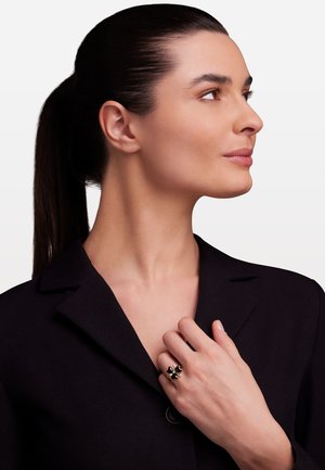 Schwarzer Blazer mit einem Reverskragen, getragen mit einer Hand, die auf dem Kragen ruht. Der Ring hat ein florales Design mit schwarzen und goldenen Akzenten.