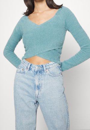 Femme portant un crop top à manches longues style cache-cœur en tissu duveteux bleu sarcelle, associé à un jean taille haute bleu clair délavé.