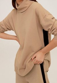 Beiger Rollkragenpullover aus weichem Strick, mit überschnittenen Schultern und schwarzen Seitenstreifen; lässige Passform mit gerippten Bündchen und Saum.