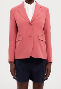 Blazer rosa con revers a ramponi, chiusura a due bottoni, due tasche frontali e una tasca sul petto. Indossato sopra una camicia bianca e pantaloni corti scuri.