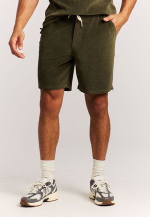 Olijfgroene shorts van zachte stof met zijzakken, een tailleband met trekkoord en een losse pasvorm, gecombineerd met grijze sportschoenen en witte sokken.