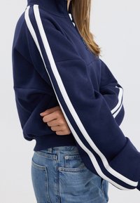 Sudadera azul marino con cremallera hasta el cuarto, con rayas laterales blancas, corte relajado y puños acanalados. Combinada con vaqueros de mezclilla de color azul claro.