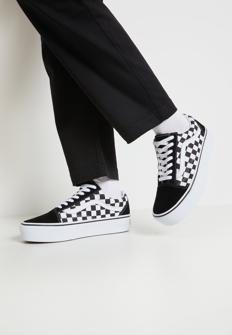 Personne portant un pantalon noir, des chaussettes blanches, des baskets à damier noir et blanc avec des lacets blancs, levant un pied sur un fond uni.