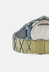 Orologio in metallo con bracciale in argento e tono oro, caratterizzato da maglie lucide e una cassa resistente. Retro contrassegnato con "DIESEL". Elementi di design classico.