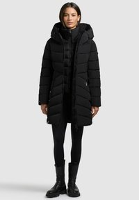 Svart puffjakke med hette, quiltet design, frontglidelås og to sidelommer. Båret over sorte leggings og robuste svarte støvler.