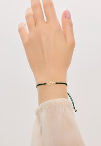 Akcent w kształcie złotego liścia na zielonej bransoletce z cordu, regulowanej za pomocą ślizgających się węzłów. Gładka powierzchnia, minimalistyczny design, idealna do noszenia w warstwach.