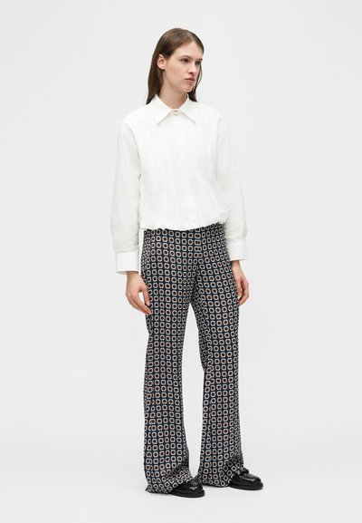 Femme portant un chemisier blanc à col et manches longues, ainsi qu'un pantalon large noir à motifs, debout avec une expression neutre.