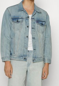 Ljusblå denimjacka med knappstängning framtill, två bröstfickor och sidfickor. Klassisk design med synliga sömmar och slitna detaljer.