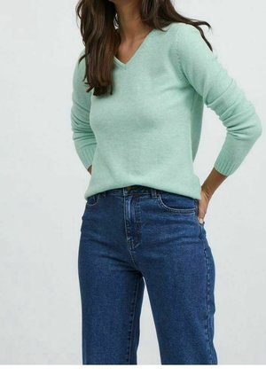 Vrouw draagt een lichtgroene V-hals trui en een high-waisted blauwe jeans, met handen op de heupen tegen een effen achtergrond.
