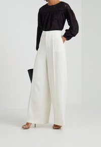 Blouse en œillets noire avec des manches bouffantes, associée à un pantalon large taille haute blanc et un sac à main noir. Des sandales à talons beiges complètent le look.