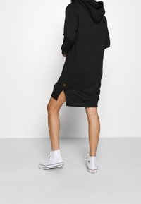 Osoba oblečená v černých šatech s kapucí a bočním rozparkem, v bílých vysokých Converse teniskách, stojící na šedé podlaze proti bílé zdi.