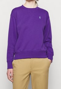 Person klädd i en lila långärmad tröja med rund halsringning och en liten blå logotyp på bröstet samt beige byxor, med händerna avslappnat vid sidorna.