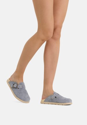Legs wearing blue slip-on clogs with small studs and an adjustable buckle on a light sole against a plain background.