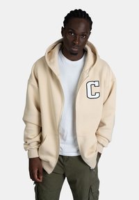 Felpa con zip beige con ampie tasche frontali e una lettera "C" ricamata in bianco sul lato sinistro, indossata sopra una maglietta bianca e pantaloni cargo verdi.