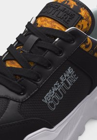 Μαύρο αθλητικό παπούτσι Versace Jeans Couture με μαύρα κορδόνια, λευκή σόλα και κίτρινη γλωττίδα με σχέδιο που φέρει το λογότυπο της μάρκας στο πλάι και στη γλωττίδα.