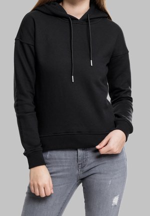Kvinde iført sort hættetrøje med snore og lysegrå revnede jeans, stående foran en ensfarvet grå baggrund.