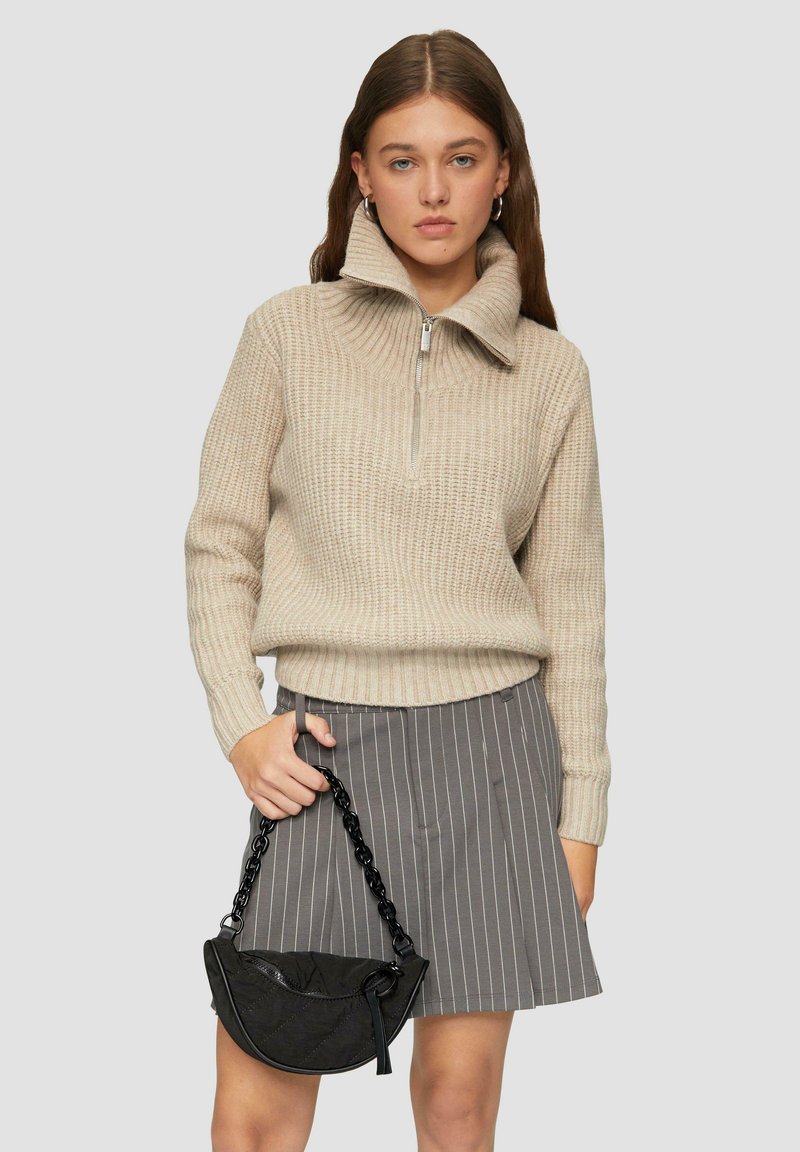 Beżowy sweter dzianinowy z wysokim kołnierzem i detalem na suwak, w połączeniu z szarym spódnicą w paski. Modelka trzyma małą czarną torbę z łańcuszkowym paskiem.