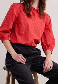 Camicia rossa con bottoni, con pois bianchi, maniche arricciate e vestibilità comoda, indossata con pantaloni in velluto a coste neri.