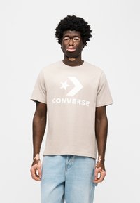 Μπεζ βαμβακερό μπλουζάκι με κοντά μανίκια, με μεγάλο λευκό αστέρι και βέλος λογότυπο, και τυπωμένο το κείμενο "CONVERSE" στο μπροστινό μέρος.