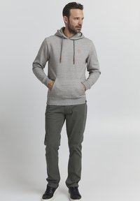 Grauer Kapuzenpullover mit verstellbaren Kordeln und Kängurutasche, kombiniert mit grünen Hosen und schwarzen Schuhen; strukturierter Stoff mit dezentem Muster.