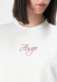 Nainen, jolla on päällään valkoinen T-paita, jossa on vaaleanpunaisella kursiivilla kirjoitettu "Hugo" -logo rinnan kohdalla. Paitaa on lyhennetty niin, että näkyy kaula ja ylävartalo.