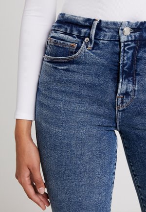 Nahaufnahme einer Person, die hoch taillierte blaue Denim-Jeans und ein weißes geripptes Langarmoberteil trägt, Hand ruht seitlich.