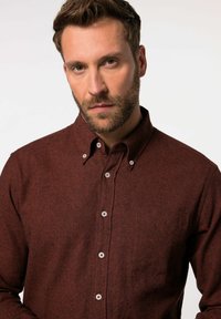 Camisa marrón de botones confeccionada con tejido texturizado, que presenta un cuello redondeado, botones blancos y un único bolsillo en el pecho.