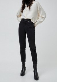 Jeans negros de talle alto con una silueta ajustada, hechos de mezclilla, combinados con una camisa de botones satinada color crema y botas negras de tobillo.