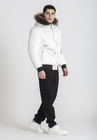 Giacca puffo bianca con cappuccio foderato in pelliccia, chiusura con zip frontale e una tasca con zip, abbinata a pantaloni della tuta neri e sneakers bianche.