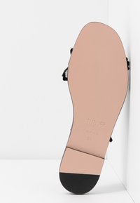 Semelle d'une sandale plate noire taille 36 avec une semelle lisse beige et noire, reposant verticalement contre un mur d'angle blanc.