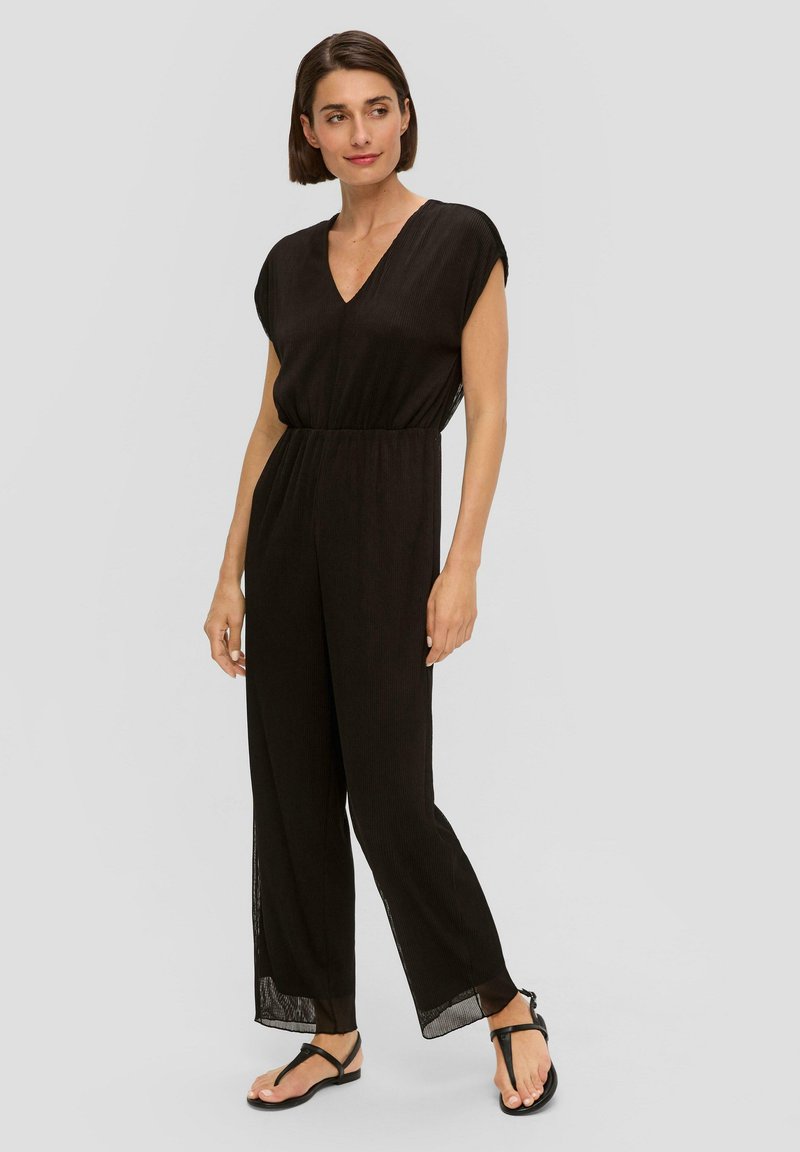 Zwarte, geribbelde jumpsuit met V-hals, korte mouwen en elastische taille. Wijde pijpen met zijsplitten. Draagt met zwarte flip-flops.
