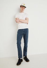 Camiseta branca de manga curta, jeans azuis, sapatos pretos com costura amarela e um boné bege com um gráfico. Posição em pé contra um fundo simples.