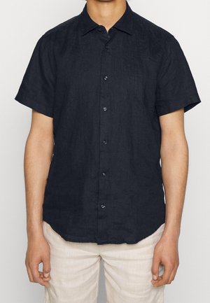 Mężczyzna w czarnej koszuli na krótki rękaw z guzikami i jasnych spodniach, stoi ze zrelaksowanymi ramionami wzdłuż ciała na jednolitym tle.