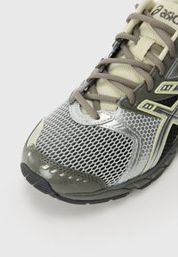 Baskets de sport grises et argentées avec des panneaux en maille, des superpositions synthétiques et une semelle en caoutchouc. Caractéristiques de fermeture à lacets et accents texturés.