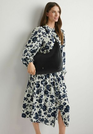 Kobieta w białej sukience z granatowym kwiatowym wzorem, trzymająca czarną torebkę na ramię, stojąca na tle jasnej ściany, patrząca w prawo i uśmiechająca się.