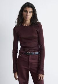 Burgunderfarbener Langarmoberteil mit Rippstruktur und ausgefranstem Saum, kombiniert mit passender burgunderfarbener Hose und einem schwarzen Gürtel mit einer goldenen Schnalle.