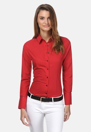 Camicia rossa con bottoni neri, maniche lunghe e vestibilità slim, abbinata a pantaloni bianchi e una cintura nera. Tessuto con una texture liscia.