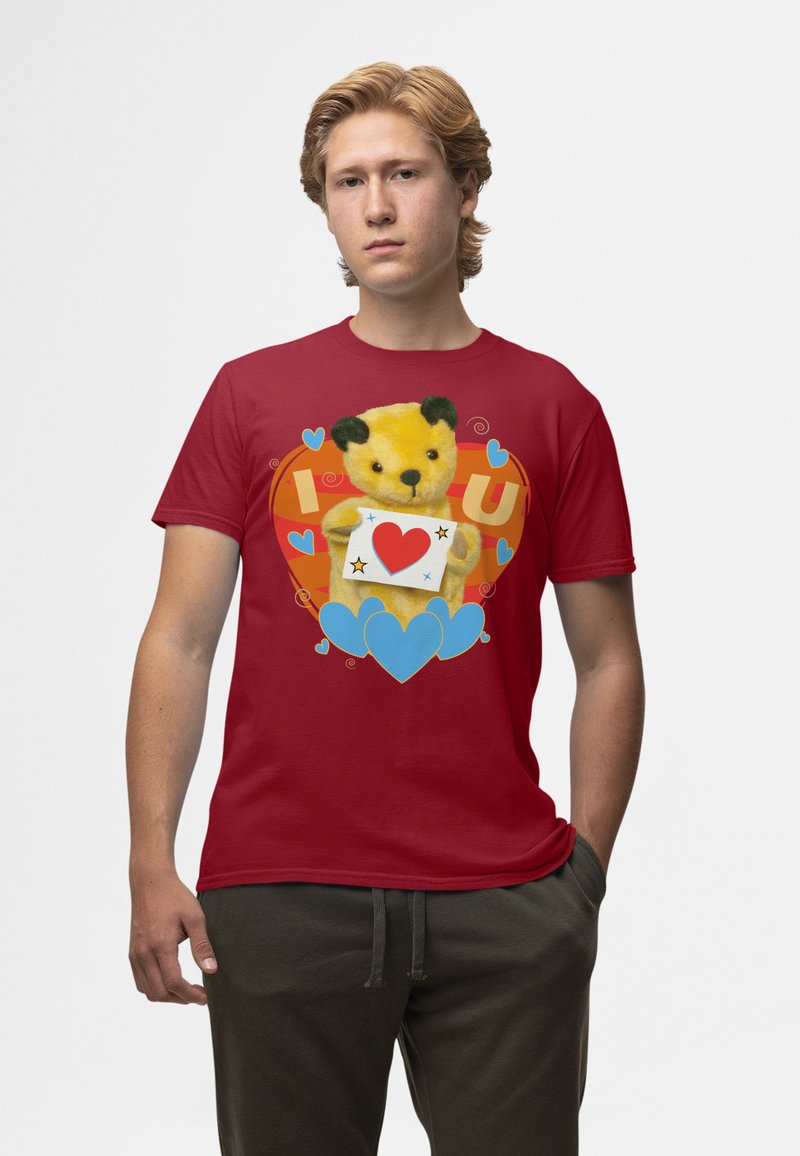 Rød T-shirt med et gult teddy bear grafik med sorte ører, der holder et hjertekort, mod en baggrund af farverige hjerter og svulster.