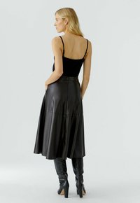 Blonde vrouw in een zwart mouwloos topje, hoge taille zwarte leren midi-rokje en zwarte kniehoog hakken, staand in zijaanzicht.