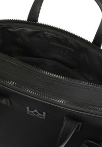 Crna torba s glatkom kožnom vanjštinom i teksturiranom tkaninom unutar. Ima zatvaranje na zip i unutarnji detalj s logotipom.