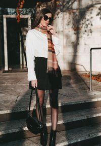 White collared shirt, black pleated skirt, and plaid scarf with brown, beige, and orange tones. Black handbag and ankle boots complete the look.