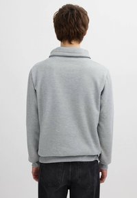 Sudadera gris ligera con cuello alto y mangas largas, que presenta una textura suave y un ajuste relajado. Combinada con jeans oscuros.