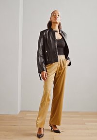 Zwarte leren jas, zwarte kanten top, tan geplooide broek en zwarte hakken. De outfit heeft gladde texturen en gestructureerde lijnen.