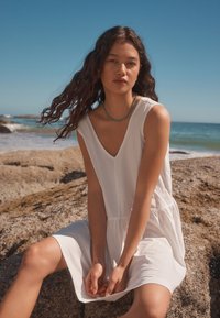 Witte, mouwloze jurk met V-hals en geplooide taille, gemaakt van zachte stof. Persoon zittend op een rots bij het strand, natuurlijk licht.