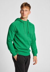 Grüne Kapuzenjacke aus weichem Stoff, mit Kordelzug-Kapuze, gerippten Bündchen und Saum, getragen mit schwarzen Hosen. Einfaches, sportliches Design.