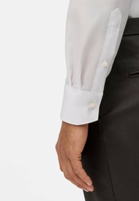 Camicia bianca a maniche lunghe con texture a tessuto semplice, polsini con bottoni e vestibilità sagomata, abbinata a pantaloni scuri.