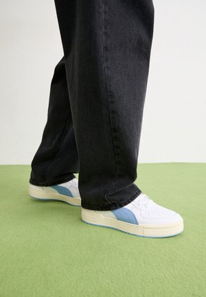 Vita sneakers med blå sidopaneler, texturerad läderöverdel och tonade sömmar. Kombinerade med mörka jeans, placerade på en grön yta.