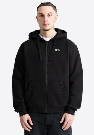 Mann mit geflochtenem Haar trägt schwarzen Hoodie mit Reißverschluss, Löwenemblem und Vordertaschen, steht vor einfachem hellem Hintergrund.