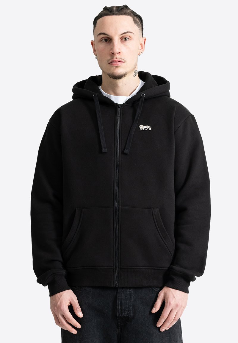Homme aux cheveux tressés portant un sweat à capuche noir zippé avec un emblème de lion et des poches devant, debout devant un fond clair uni.