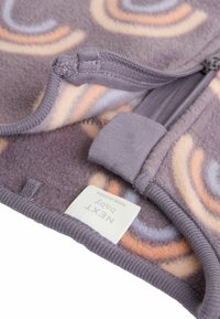 Pilkas fliso audinys su daugiakosėmis spiralinėmis formomis, užsegamas užtrauktuku, su etikete "NEXT BABY." Minkšta tekstūra ir apvalus dizainas.